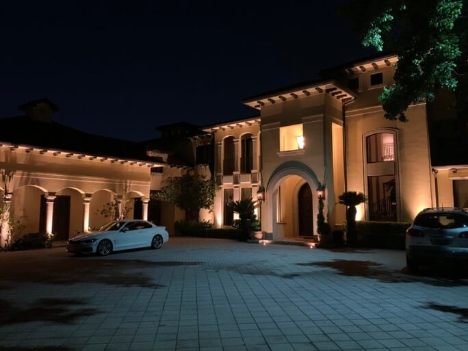 The memorial villages tx villa lit up at night with an archway to the front door and a lit up window above it. There are two cars in the driveway.