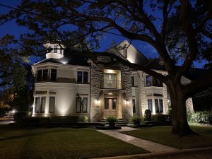 The 3 story Bellaire Tx abode sits lit up at night with a large tree to the right of the walkway