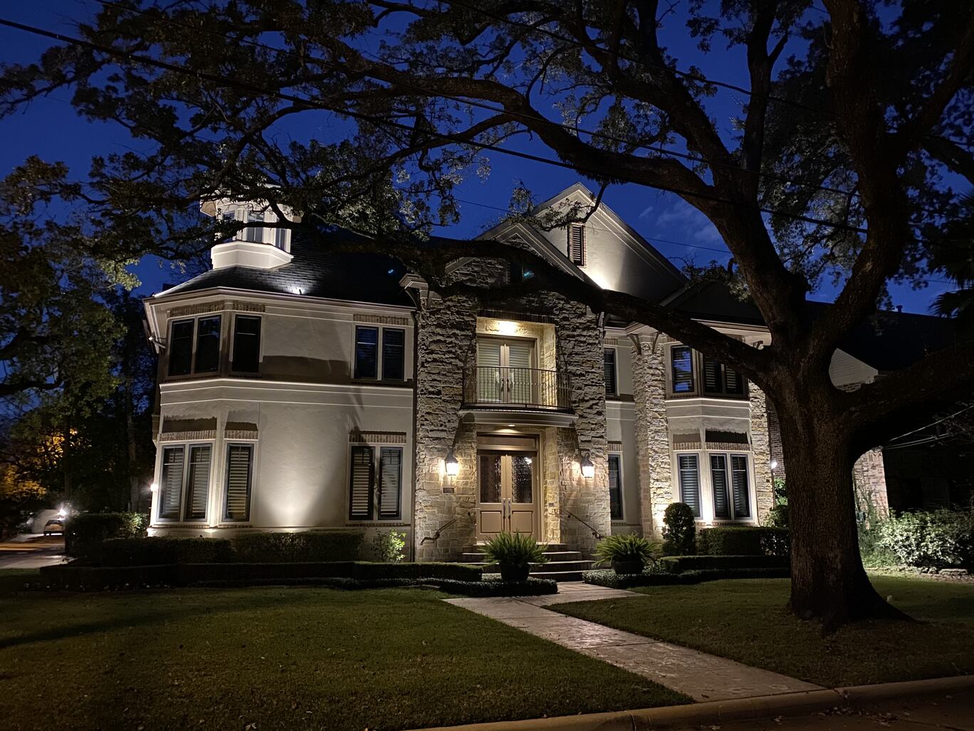 The 3 story Bellaire Tx abode sits lit up at night with a large tree to the right of the walkway