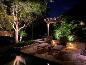 Two pool chairs sit under a pergola illuminated by deck lighting at night
