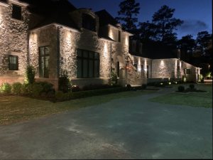 A stone two story house with a large driveway in front is lit up at night with downlighting
