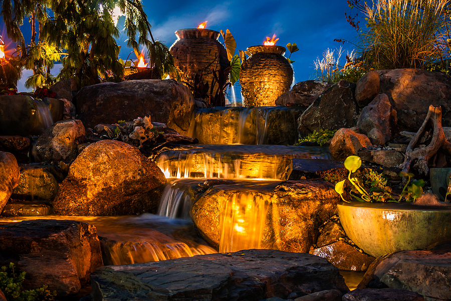 A backyard brook with stones, jugs, and small waterfalls illuminated by waterproof lighting leads to a magic garden. 