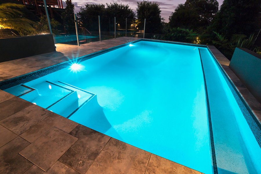 Blue water swimming pool with flashing lights and tiled flooring, set in a garden surrounded by dark trees at night.