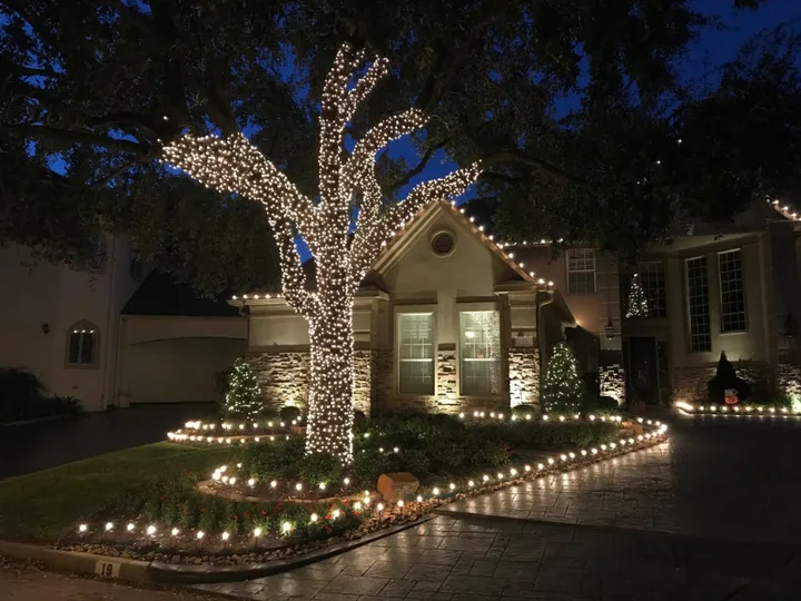 Holiday lighting across the roofline of a home with a mature oak wrapped in warm white lights