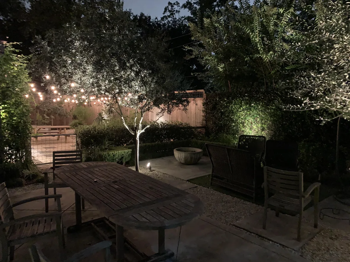 hl-res-backyard-patio-1180.webp Patio and garden seating area at night with string lights overhead and lit planting beds